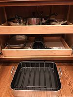 Wide view of two open kitchen drawers containing various cookware including roasting pan, saucepans with lids, kitchen clock, strainer basket.