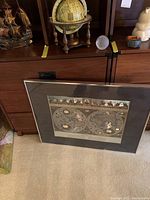 Photo showing the framed antique-style world map print leaning against furniture, with the wooden desk globe and part of the glass globe visible on top of a wooden cabinet.