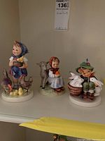 Three Hummel figurines displayed on shelf: girl with duck, girl with lamb, boy with rabbits.