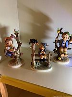 Wide shot showing all three Hummel figurines on a white surface, highlighting each figure's distinct pose and decorative elements with birds and branches.