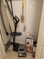 Closet floor view showing Miele canister vacuum, Shark steam mop box, Bona kit box, wall-hung manual mop and dusters, gray folded ironing board