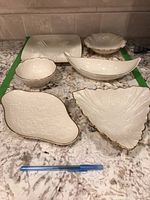 Photo showing six Lenox porcelain serving pieces of various shapes with embossed floral and shell patterns and gold trim.