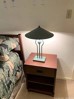 Table lamp placed on a wooden nightstand next to a bed, showing the full lamp with conical shade and rectangular base.