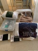 Three new pillows packaged, mattress cover, brown and cream throws, plastic storage tote containing various items including rolls visible inside, white shoe shelf box on the floor.