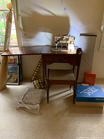 Full view of Singer sewing machine setup on a wooden sewing cabinet with matching stool, accompanied by sewing book and box of accessories on floor.