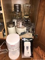 Photo showing assortment of small kitchen appliances inside wooden cabinet including Osterizer blender, Cuisinart food processor and mini prep, white cylindrical grinder, Sunbeam Hot Shot dispenser, various attachments and parts