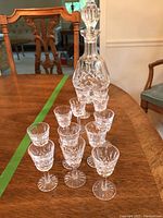 Decanter with stopper at back and 12 cordial glasses arranged in front on wooden table