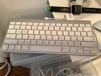 Apple wireless keyboard on top of plastic container with charging cables and Apple Watch next to it.