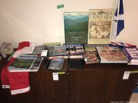 Photo showing various books and DVDs with Canadian, Chinese, Scottish history and travel themes laid out on a dark-colored wooden cabinet, partially draped with a Canadian flag.