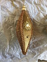 Close-up image showing front side of tear-drop shaped ornament in gold fabric with copper trim and gold embroidery with beads.