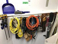 IRWIN Quick-Grip clamps in yellow and blue hanging on wall with extension cords and work light cords nearby, with broom and dustpan above them.