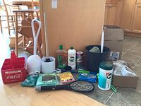 Photo showing various cleaning tools and supplies including plungers, brushes, cleaning liquids, wipes, cloths, bucket and a red Coca-Cola container.