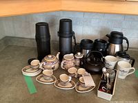 Overview of the entire espresso set showing the handmade ceramic tea pot, cups, sugar bowl, creamer, and all accessories including black insulated mugs, thermoses, white mugs, metal kettle, spoons and tea strainers arranged on a counter.