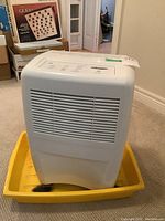 Front and side views of the white Whirlpool dehumidifier placed inside a yellow plastic tray on carpeted floor.