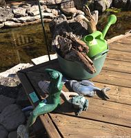 Full lot view on wooden deck beside pond, showing bucket with driftwood, watering can inside bucket, two fish figures, one frog, and mermaid figure