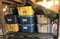Seven plastic storage bins stacked on shelving, each labeled for garden supplies like fertilizer, tarps, pots, grass seed, and ropes, covered partially with a green tarp