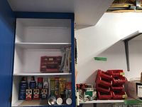 Shelf with various cans, bottles, and small red organizer box inside a blue cabinet.