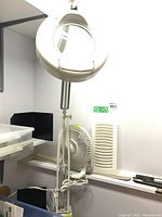 Full view of the clip-on magnifying lamp clamped to a surface alongside the Holmes table fan on a shelf.