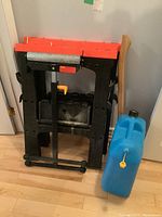 Black WorkForce saw horse with orange extender, blue plastic 5 gallon water jug, and wooden handled axe leaning against wall.