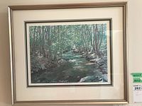 Framed print showing a colorful stream flowing through trees, visible signature and number on bottom margin.