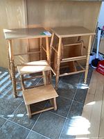 Two light-colored wooden step stools with retractable steps, placed on tiled floor against wooden cabinetry, showing front and side views.