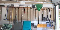 Wide view of multiple rakes, shovels, pitchforks, spades, and snow shovels hanging on the wall of a garage.
