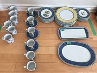 Overview of all pieces: white mugs, cups with saucers, plates, and two platters on hardwood floor