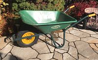 Full side view of green Tufx wheelbarrow showing metal frame and yellow plastic wheel on stone patio.