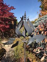 Full view of tall metal garden windmill showing stylized curved sails and rusted gold paint, set outdoors in garden surrounded by plants.