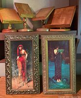 Four wooden book stands displayed on a shelf with two vertically oriented framed reproduction prints of classical art featuring female figures. The prints have ornate frames.