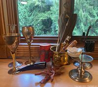 Set of chalices, candlesticks, ritual tools including brushes, wands, black blown horn, brass cauldron, small dishes, and red decorative container arranged on wooden surface in front of window.
