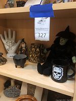 Shelf containing multiple items including a ceramic hand sculpture with palmistry lines, a wizard figure, bronze serpent sculpture, black cauldron bowl, skull mug, and a small plaque.