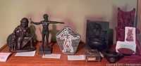 Wide shot showing seven assorted figurines and statues on wooden surface, including kneeling male figure, standing male figure, pentagonal decor plaque, small bowl, metal box, seated figure, and tall red statue.