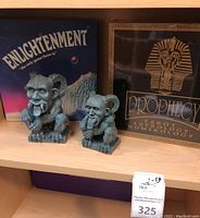Two gargoyle figurines in front of Prophecy and Enlightenment board game boxes on a wooden shelf.
