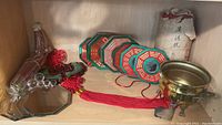 Shelf containing assorted Asian inspired decor: brass bowl with tripod base, octagonal bagua plaques, red yarn hanging decorations with green fish pendants, small mirrors, and a wrapped tablecloth.