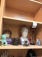 Shelf view showing the full lot including phrenology head statue, Lady swimmer candle holder, horned god statue, gargoyle snow globe, glow-in-the-dark alien figure, St. Brigid's Cross plaque, Paul Bunyan collectible, and other small decorative items.