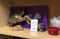 Photo showing the Tree-Woman sculpture lying horizontally on shelf next to purple Mastery game box and small ceramic bowls