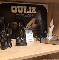 Shelf showing the Ouija board box, two mermaid black sculptural candle holders, and a lighted candle décor with electric flame and power cord.