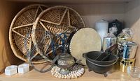 Wiccan ritual items arranged on wooden shelf including woven rattan trays with pentagram design, hand drum, various candles and holders, cast iron cauldron, ornate incense burner, and other accessories.