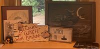 Photo showing small framed witch-themed artwork, wooden signs with witch-related phrases, and small witch figurines arranged on a wooden surface in front of a window.