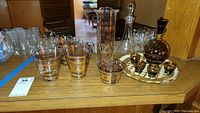 Photo of the full barware set on wooden surface including pitcher, two decanters, six bar glasses with gold designs, six small amber glasses, and clear tray.