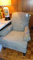 Armchair in a room setting with tags attached to upper backrest showing script patterned upholstery and wooden legs.