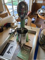 Photo shows the Hamilton Beach DrinkMaster with green stand, stainless steel mixing cup, manual, and recipe book on a metal tray in a room with boxes and a window.
