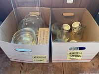 Two cardboard boxes containing assorted clear glass Pyrex bowls with lids, clear glass cups, small pitcher, and white ceramic ramekins.