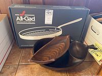 All-Clad stainless steel non-stick grille pan in original box with wooden serving bowl and spoon and ceramic dish on a tiled surface.