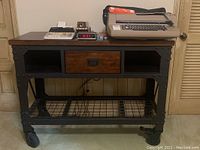 Front view of industrial workbench with wooden top, central drawer, and metal frame on caster wheels. Items placed on top include an electric calculator, clock, typewriter, and small organizer.