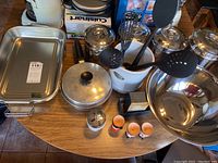 Photo showing a variety of stainless steel cookware including pots, pans, mixing bowl, and various kitchen utensils stored in a white ceramic crock with additional small kitchen items on the table.