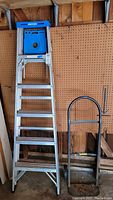 Full view of assembled 6ft aluminum Werner ladder next to a black metal dolly. Ladder has blue plastic top with Werner label and multiple steps with locking braces.