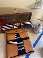 Wide shot showing wooden cutlery case with knives and forks, leather pouch with cutlery, and metal teapot on kitchen counter.
