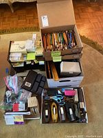 View of multiple boxes filled with various office supplies including pencils, pens, tape dispensers, staplers, scissors and other small desk items.